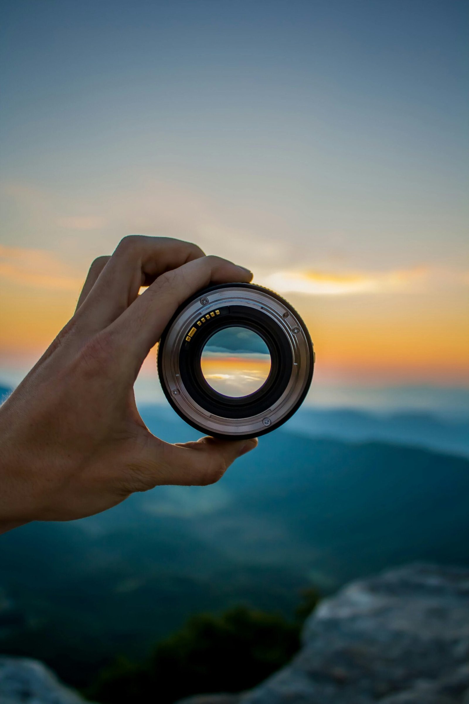A hand holding a camera lens against a scenic sunset background, capturing nature's beauty.